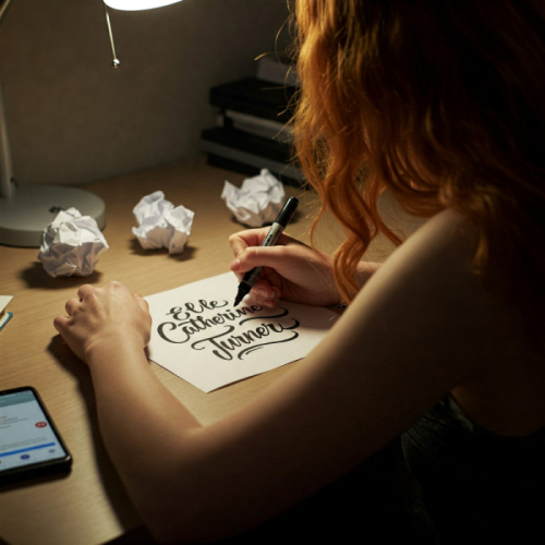A person with red hair sits at a desk and writes the name "Elle Catherine Turner" in decorative script on a piece of paper. Crumpled papers and a phone are on the desk.